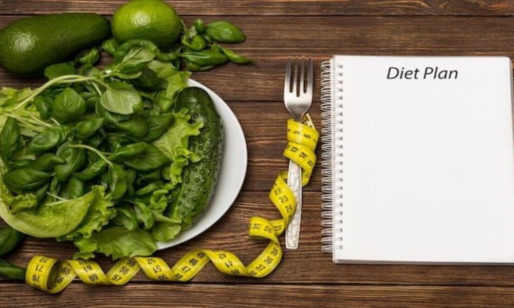 Piring berisi sayuran hijau, meteran jahit kuning, garpu, dan buku catatan bertuliskan 'Diet Plan' di atas meja kayu.