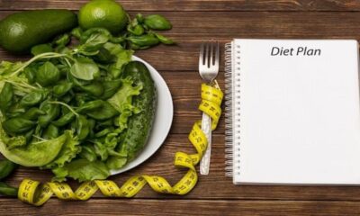 Piring berisi sayuran hijau, meteran jahit kuning, garpu, dan buku catatan bertuliskan 'Diet Plan' di atas meja kayu.