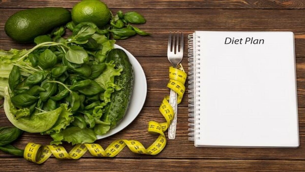 Piring berisi sayuran hijau, meteran jahit kuning, garpu, dan buku catatan bertuliskan 'Diet Plan' di atas meja kayu.