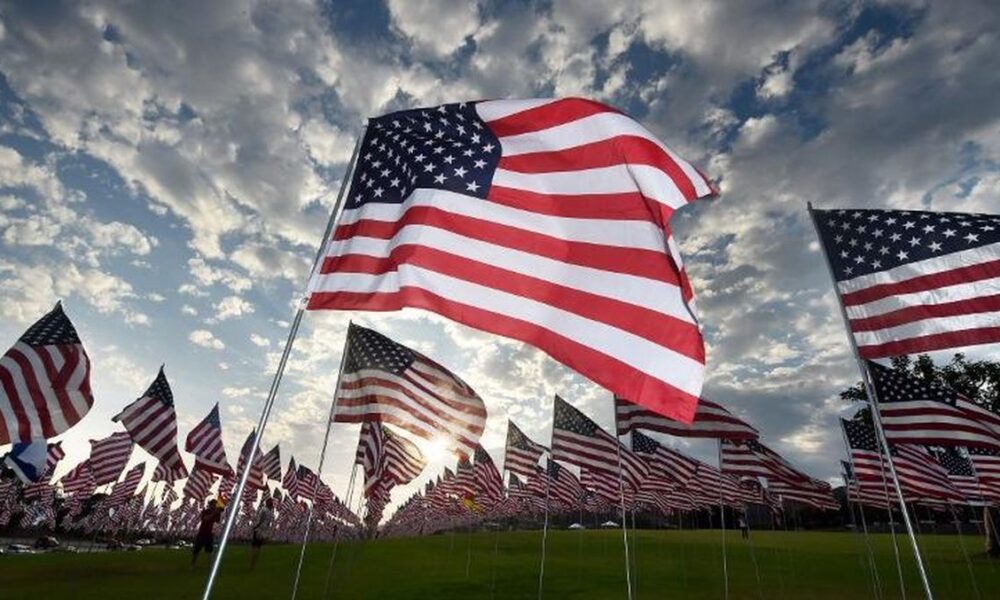 Bendera Amerika Serikat berkibar di tiang-tiang di lapangan rumput dengan latar belakang langit berawan