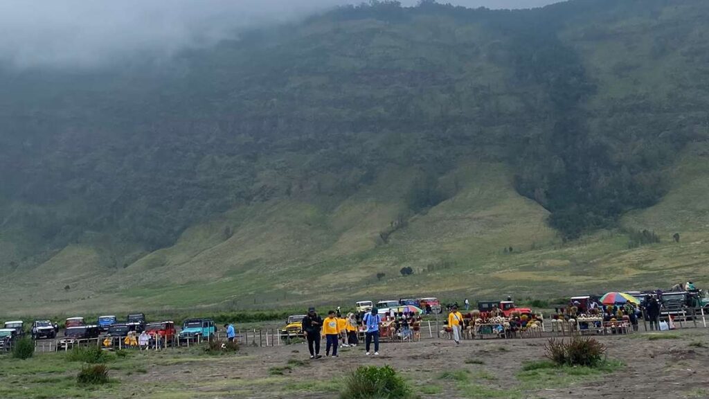 Wisata bromo kembali dibuka usai kebakaran, Sekelompok wisatawan berjalan di area terbuka dengan latar belakang tebing bukit yang diselimuti kabut tipis dan jejeran jip wisata terparkir di kejauhan