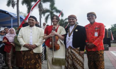 Putri Ariani memegang biola berdiri berdampingan dengan pejabat daerah mengenakan pakaian adat Jawa di luar ruangan