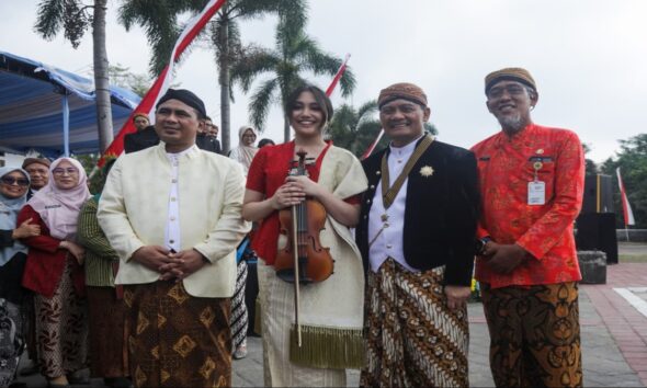Putri Ariani memegang biola berdiri berdampingan dengan pejabat daerah mengenakan pakaian adat Jawa di luar ruangan