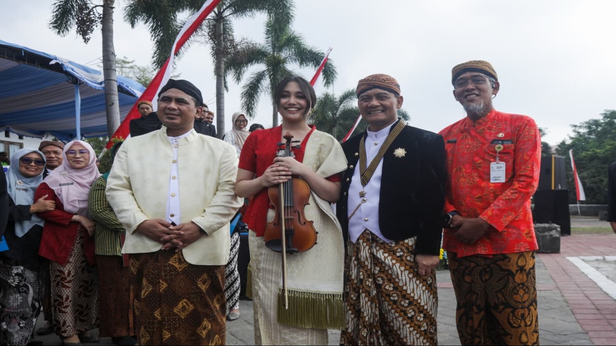 Putri Ariani memegang biola berdiri berdampingan dengan pejabat daerah mengenakan pakaian adat Jawa di luar ruangan