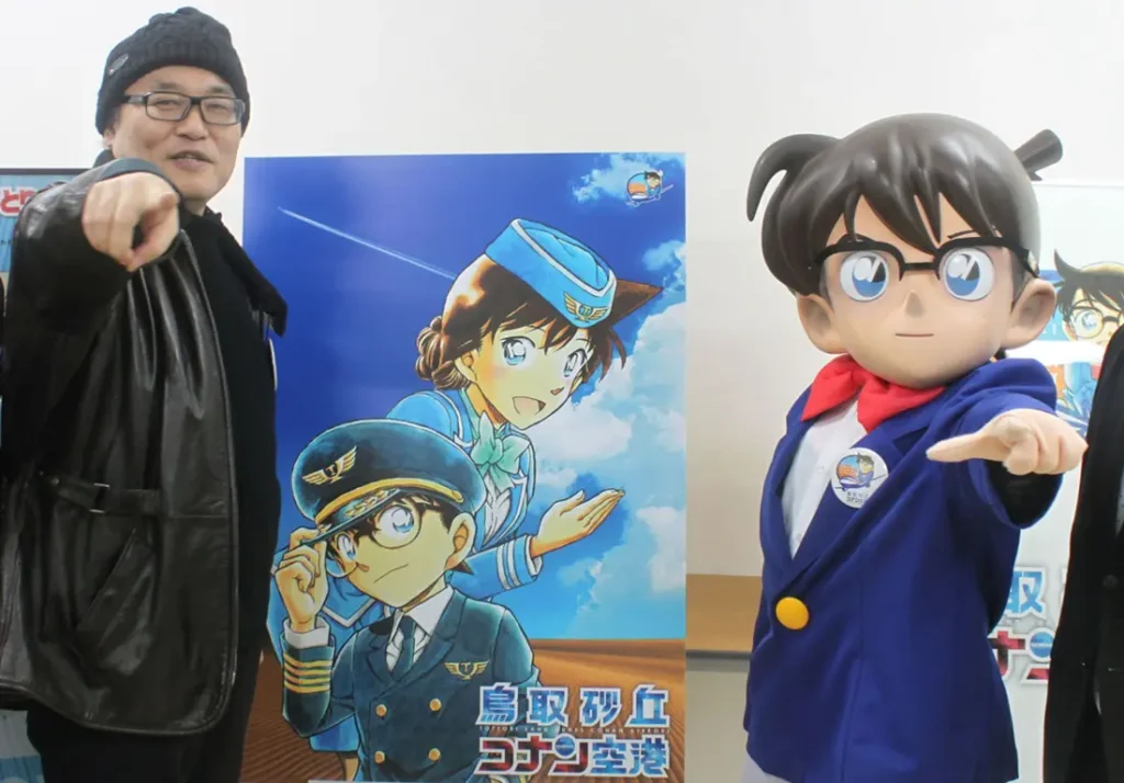 Aoyama Gosho Bocorkan 1 Rahasia Penting Akhir Cerita Detective Conan 53 Mangaka Gosho Aoyama Gosho berdiri di sebelah poster ilustrasi Conan dan Ran dalam balutan seragam pilot, ditemani karakter berkostum Conan