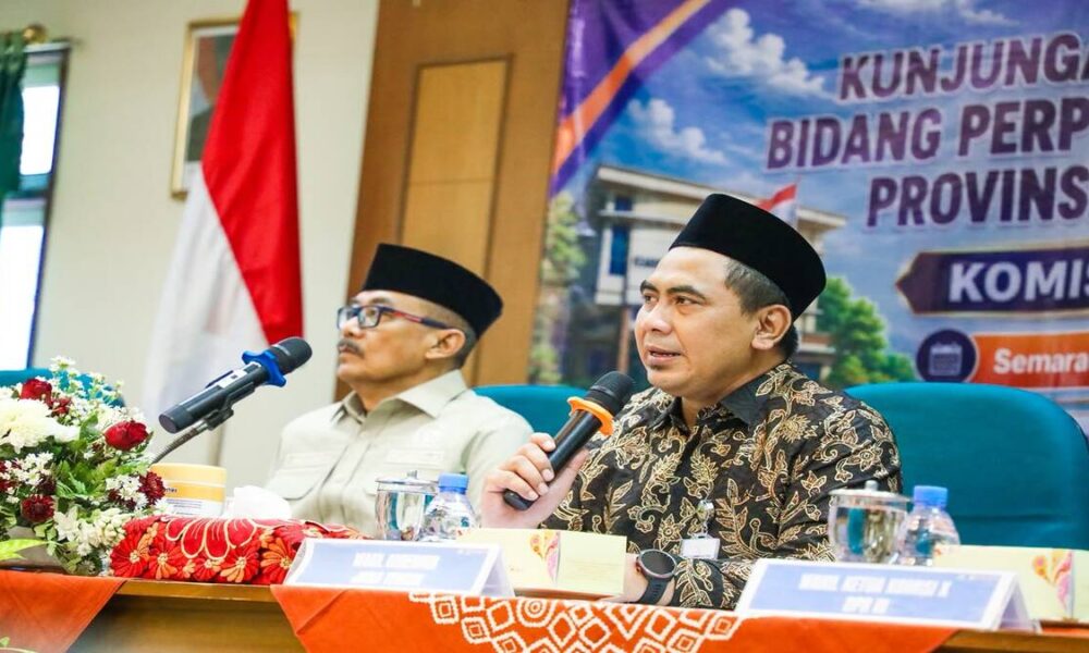 Dua pria berpeci duduk di meja konferensi, pria di depan memegang mikrofon dan berbicara