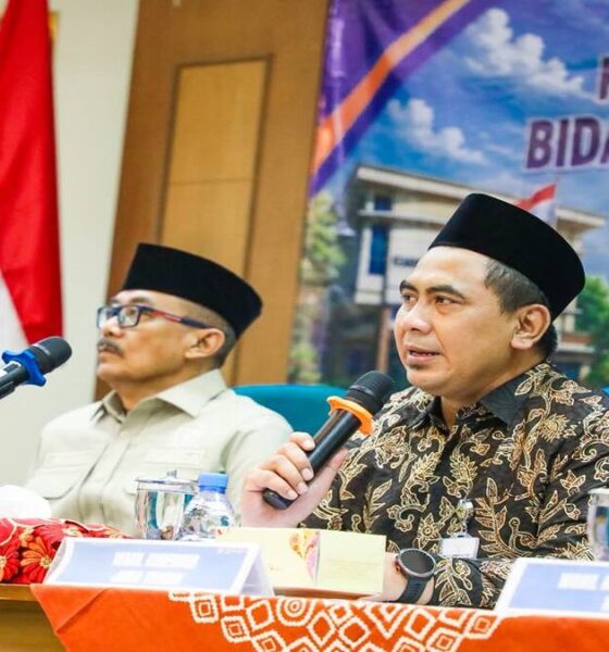 Dua pria berpeci duduk di meja konferensi, pria di depan memegang mikrofon dan berbicara