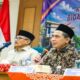 Dua pria berpeci duduk di meja konferensi, pria di depan memegang mikrofon dan berbicara