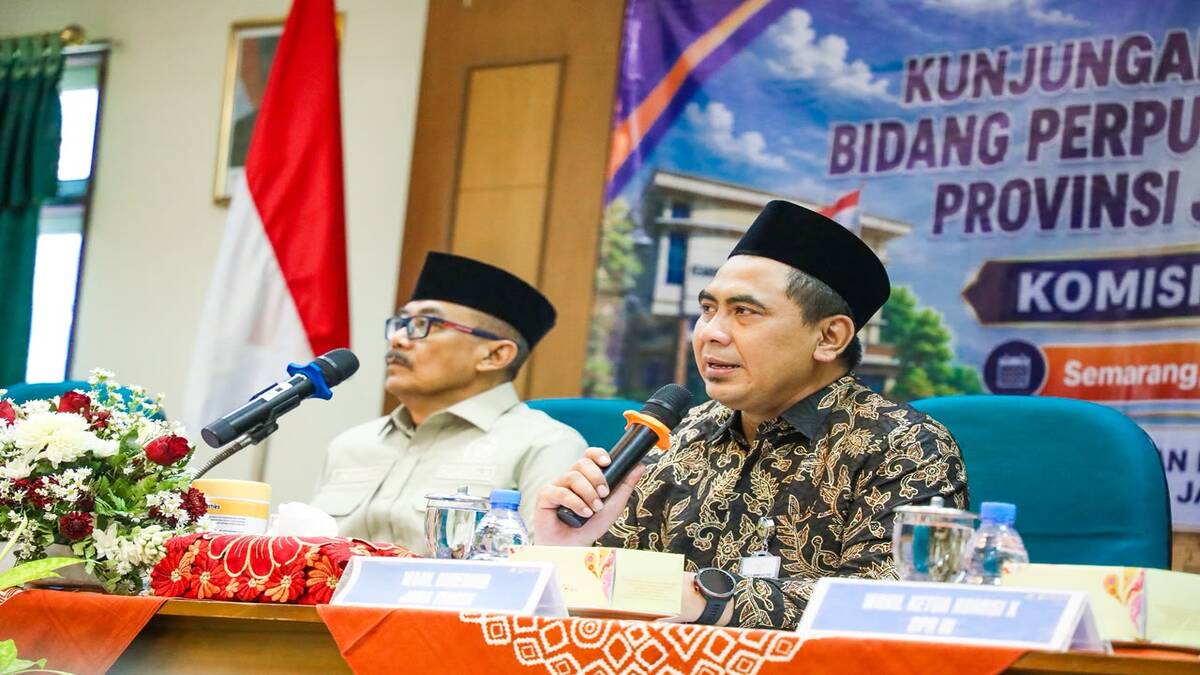 Strategi Dahsyat Pemprov Jateng untuk Tingkatkan Literasi Warga 51 Dua pria berpeci duduk di meja konferensi, pria di depan memegang mikrofon dan berbicara