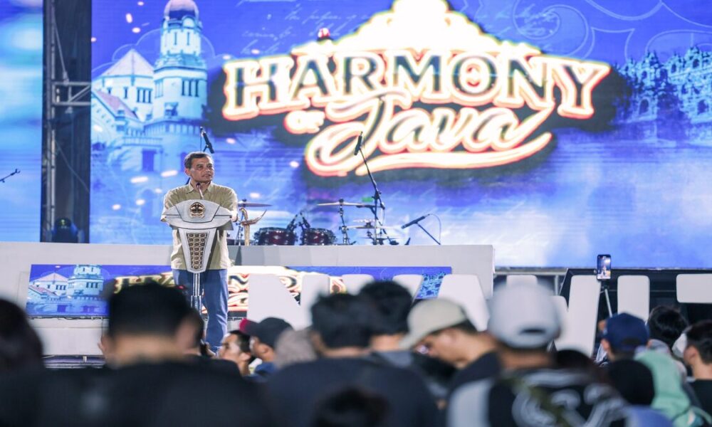 Seorang pria berdiri di atas panggung di depan layar bertuliskan Harmony of Java, berbicara di hadapan penonton yang memadati area