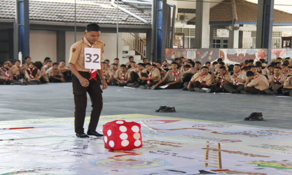 Seorang anggota Pramuka bernomor dada 32 berdiri di sebelah dadu merah besar di atas papan permainan raksasa, disaksikan oleh peserta lain yang duduk di latar belakang