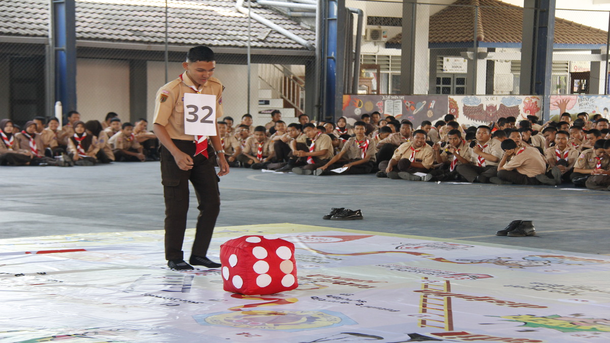 Ular Tangga Berintegritas: Inovasi Spektakuler SMKN 1 Semarang di HUT RI 51 Seorang anggota Pramuka bernomor dada 32 berdiri di sebelah dadu merah besar di atas papan permainan raksasa, disaksikan oleh peserta lain yang duduk di latar belakang