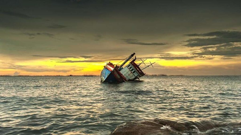 Kapal karam yang miring di perairan tenang saat senja berpendar kuning di langit berawan