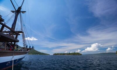 Kapal phinisi berlayar di perairan biru dengan latar belakang pulau-pulau kecil di bawah langit cerah