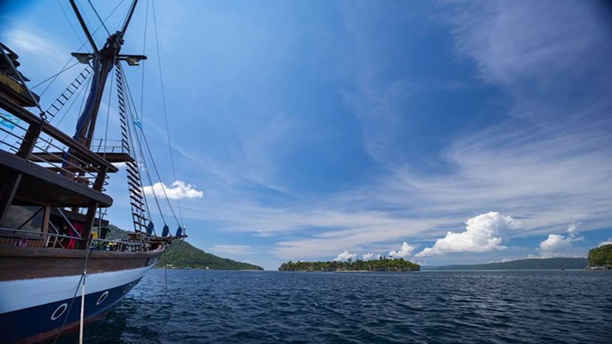 Kapal phinisi berlayar di perairan biru dengan latar belakang pulau-pulau kecil di bawah langit cerah