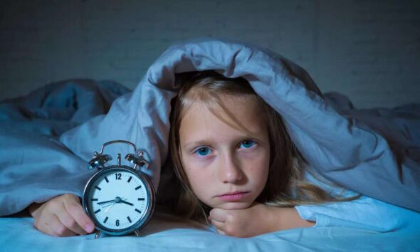 Seorang anak perempuan yang tampak kesulitan tidur berbaring di bawah selimut sambil memegang jam alarm di malam hari