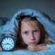 Waspada! Ini Efek Buruk Anak Kurang Tidur yang Menghancurkan Masa Depan Pertumbuhannya 55 Seorang anak perempuan yang tampak kesulitan tidur berbaring di bawah selimut sambil memegang jam alarm di malam hari