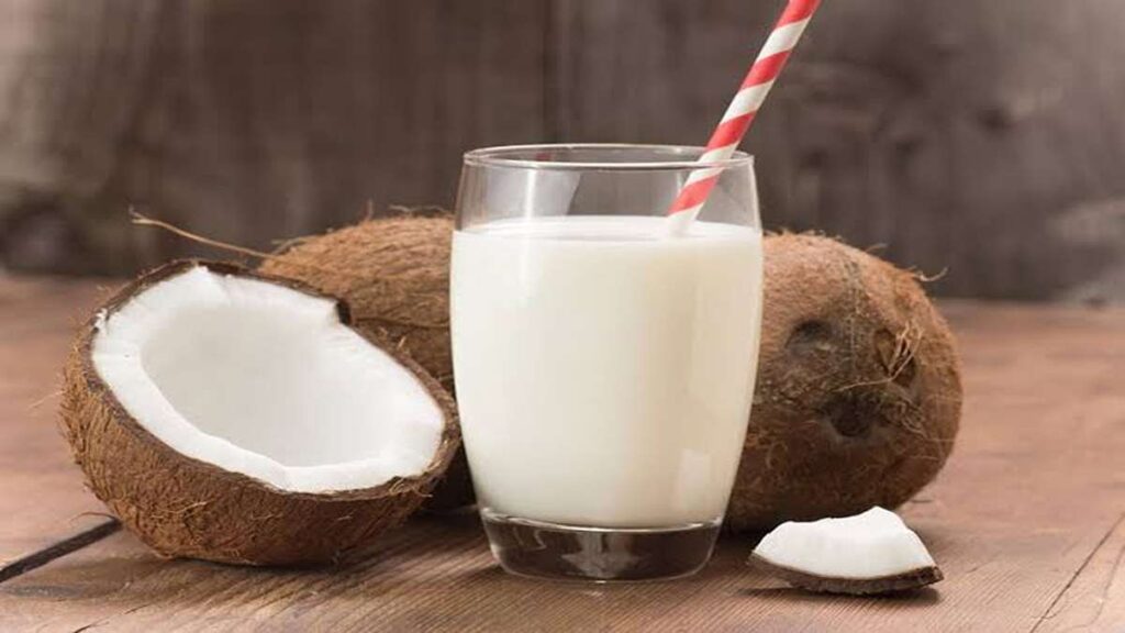 Perbandingan Jumlah Protein Berbagai Jenis Susu dari Almond hingga Susu Sapi 58 Secangkir susu kelapa putih dengan sedotan merah putih, dikelilingi oleh kelapa utuh dan potongan kelapa di atas meja kayu