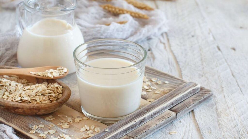 Perbandingan Jumlah Protein Berbagai Jenis Susu dari Almond hingga Susu Sapi 56 Segelas susu havermut di atas meja kayu dengan mangkuk berisi butiran havermut dan teko kaca berisi susu di latar belakang