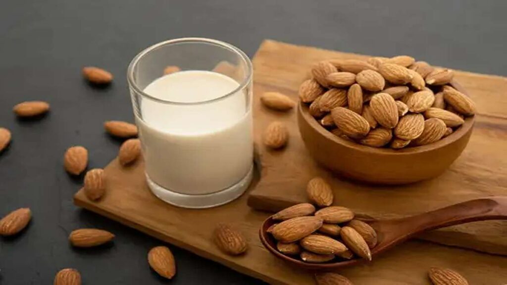 Perbandingan Jumlah Protein Berbagai Jenis Susu dari Almond hingga Susu Sapi 57 Segelas susu almond di samping mangkuk dan sendok berisi biji almond utuh di atas talenan kayu