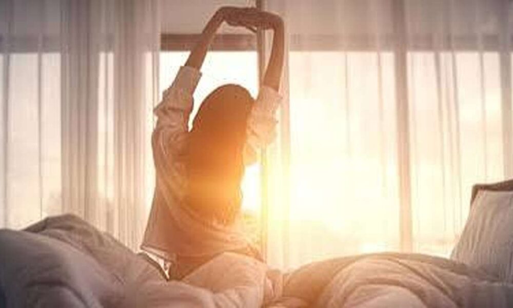 Seorang wanita meregangkan tangan di tempat tidur saat matahari terbit di dekat jendela kamar
