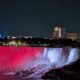 Air terjun Niagara pada malam hari diterangi cahaya merah dan putih di bawah langit malam dengan latar belakang gedung-gedung kota yang bercahaya