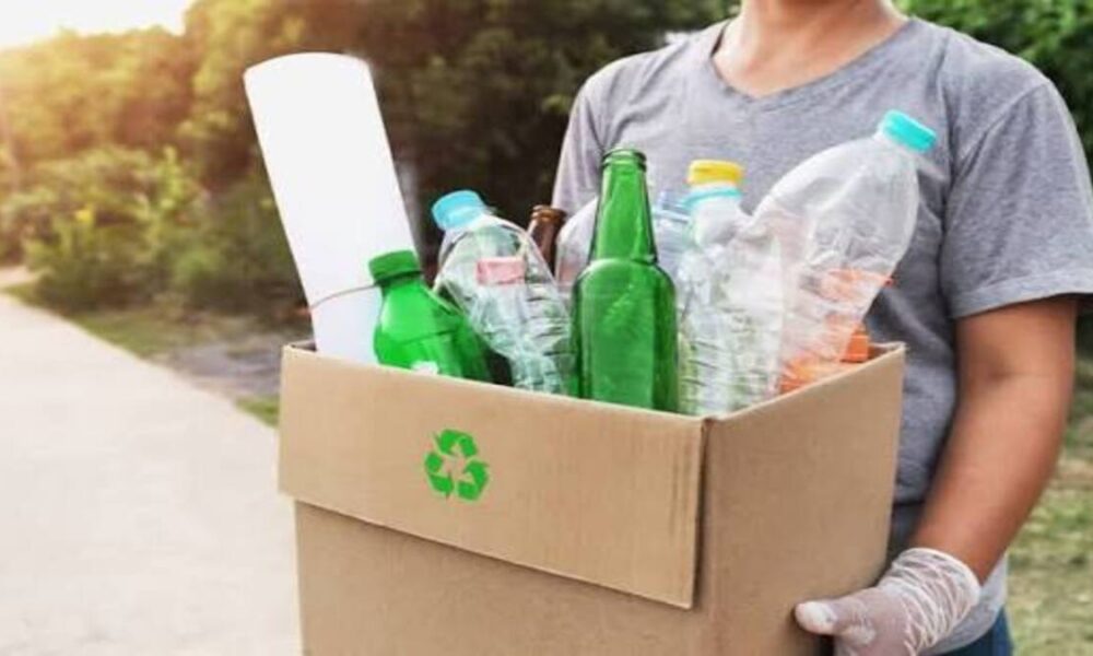 Seseorang mengenakan sarung tangan membawa kardus berisi botol plastik, botol kaca, dan gulungan kertas untuk didaur ulang