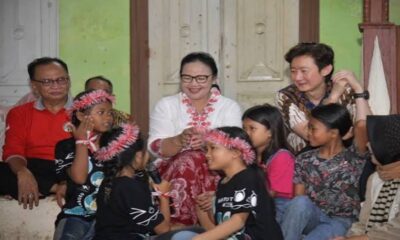 Sekelompok anak perempuan mengenakan hiasan kepala merah dan hitam sedang bernyanyi bersama orang dewasa dan tamu di ruangan