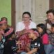 Sekelompok anak perempuan mengenakan hiasan kepala merah dan hitam sedang bernyanyi bersama orang dewasa dan tamu di ruangan
