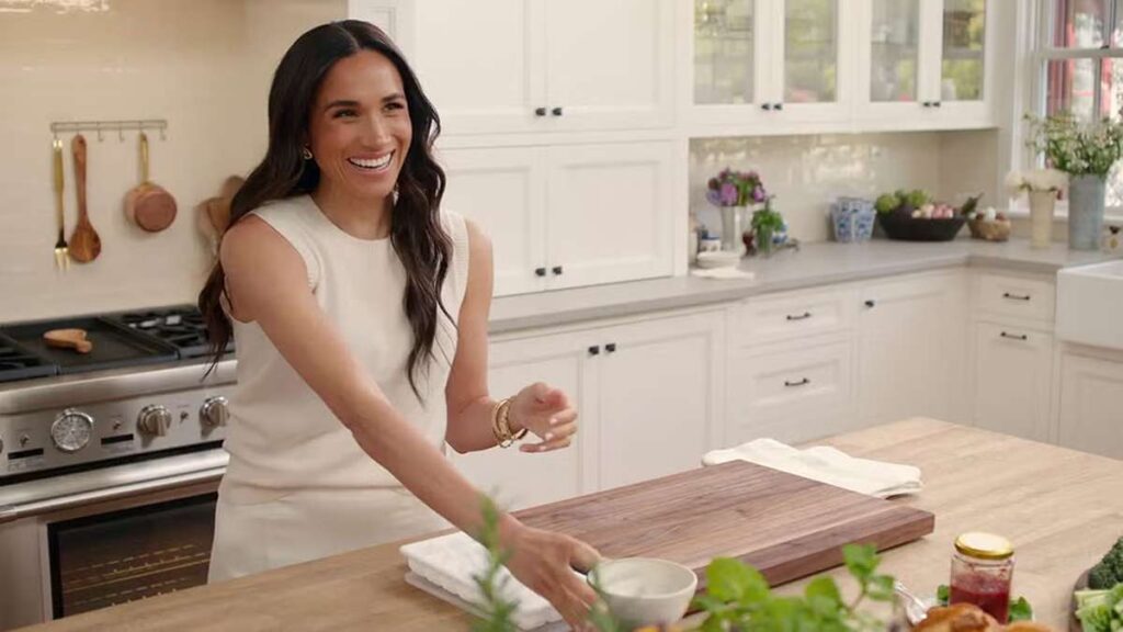 Serial Netflix Terbaru Meghan Markle kembali berkarier, Meghan Markle tersenyum di dapur putih sambil memegang mangkuk kecil di dekat meja pulau kayu