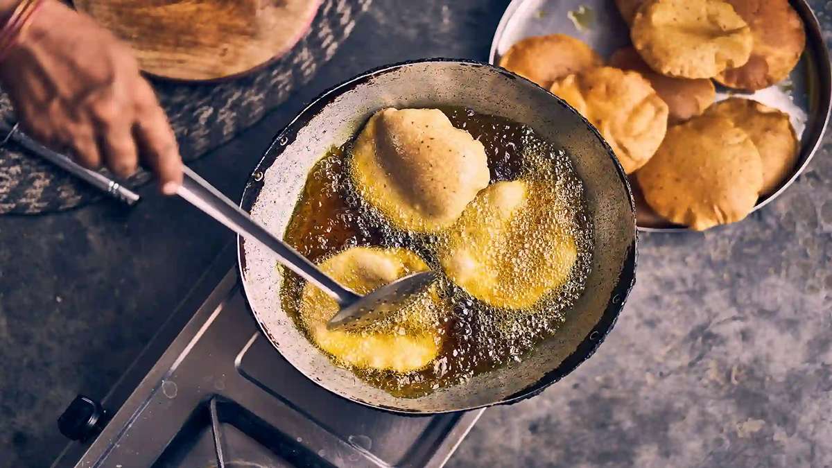 Seseorang sedang menggoreng puri di dalam wajan berisi minyak panas dengan saringan, dan piring berisi roti puri matang di sampingnya