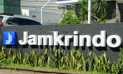 Papan nama perusahaan Jamkrindo berwarna putih dengan latar belakang dinding abu-abu dan tanaman hias di depannya