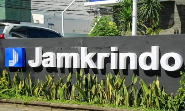 Papan nama perusahaan Jamkrindo berwarna putih dengan latar belakang dinding abu-abu dan tanaman hias di depannya