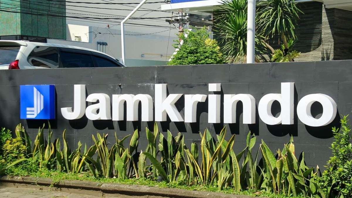 Papan nama perusahaan Jamkrindo berwarna putih dengan latar belakang dinding abu-abu dan tanaman hias di depannya