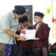 Seorang pria mengenakan peci dan sarung menyerahkan sertifikat kepada santri berseragam jas merah marun dan peci hitam