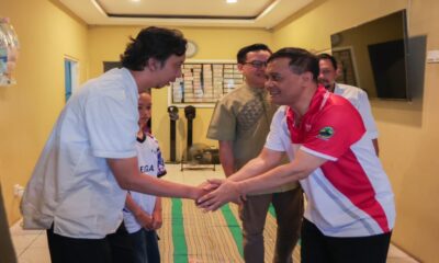 Dua pria berjabat tangan di dalam ruangan, disaksikan oleh beberapa orang di latar belakang