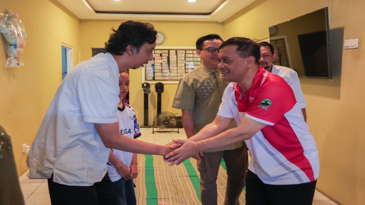 Dua pria berjabat tangan di dalam ruangan, disaksikan oleh beberapa orang di latar belakang
