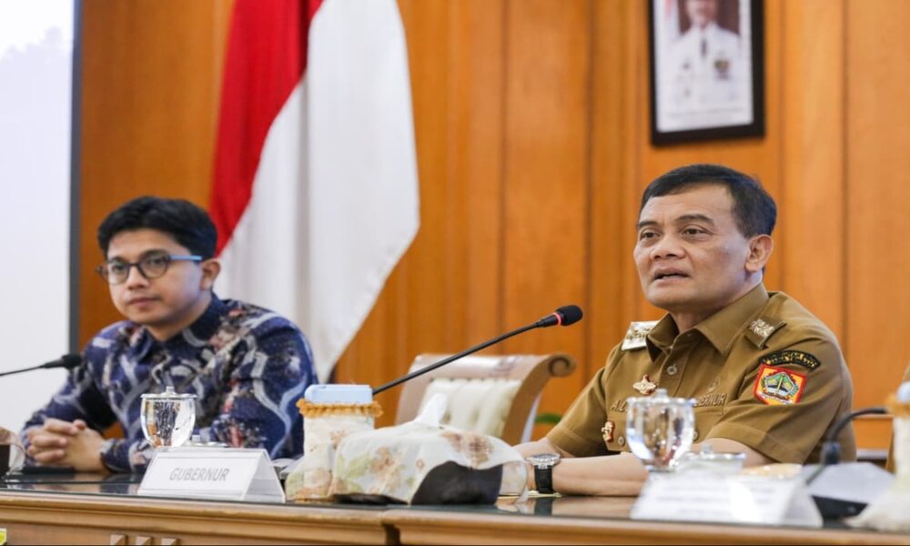 Nana Sudjana mengenakan pakaian dinas saat menghadiri rapat dengan latar belakang bendera merah putih