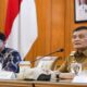 Nana Sudjana mengenakan pakaian dinas saat menghadiri rapat dengan latar belakang bendera merah putih