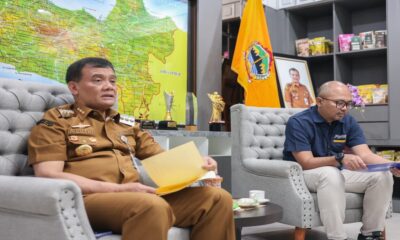 Dua orang pria sedang duduk di sofa sambil memegang dokumen di ruangan berlatar belakang peta dan rak pajangan