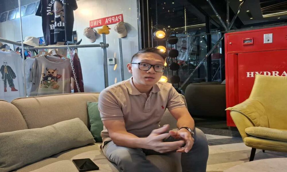 Seorang pria berkacamata duduk di sofa krem di dalam ruangan, dengan latar belakang rak pakaian dan kotak hidran merah