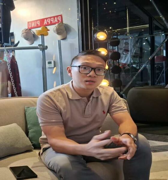 Seorang pria berkacamata duduk di sofa krem di dalam ruangan, dengan latar belakang rak pakaian dan kotak hidran merah