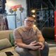 Seorang pria berkacamata duduk di sofa krem di dalam ruangan, dengan latar belakang rak pakaian dan kotak hidran merah