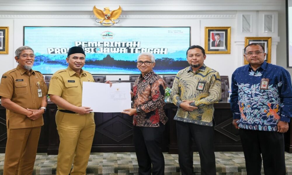 Lima pejabat berdiri berjejer di dalam ruangan, seorang pria di tengah memegang dokumen yang diserahkan kepada rekan di sebelah kirinya