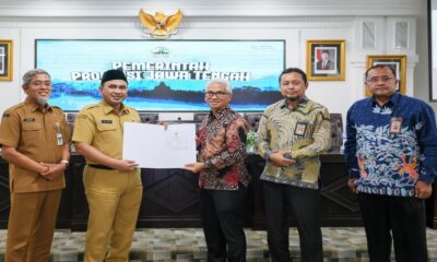 Lima pejabat berdiri berjejer di dalam ruangan, seorang pria di tengah memegang dokumen yang diserahkan kepada rekan di sebelah kirinya