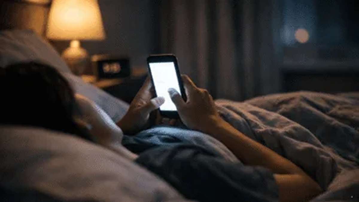 Seseorang sedang berbaring di tempat tidur di dalam kamar yang gelap sambil menatap layar ponsel yang menyala terang