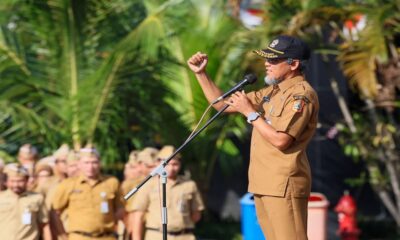 Seorang pria berseragam dinas cokelat dan bertopi menyampaikan pidato dengan gestur tangan mengepal di hadapan audiens di luar ruangan