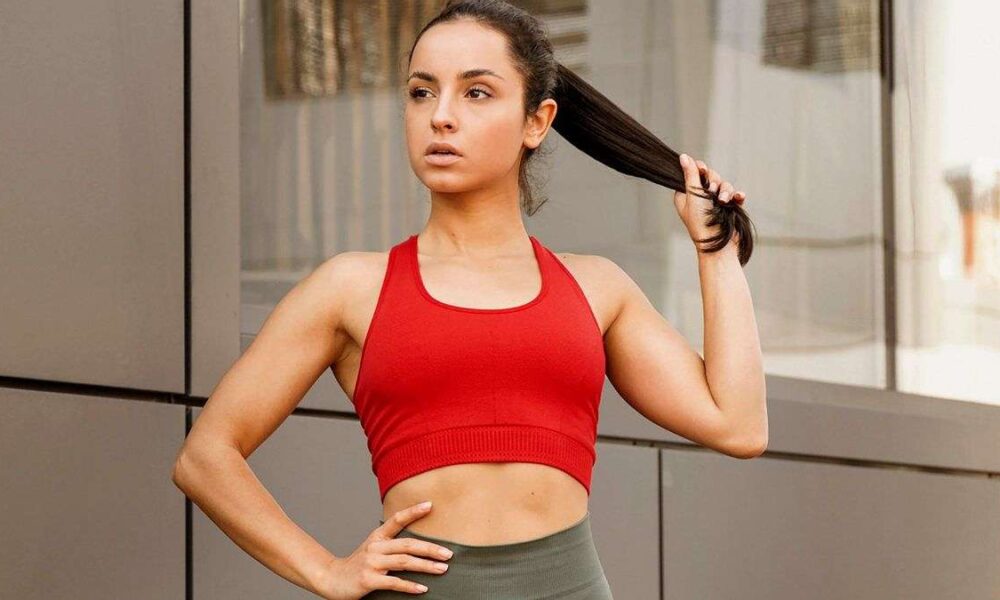 Seorang wanita berambut gelap memakai sport bra merah dan memegang rambutnya, berpose di luar ruangan dengan latar dinding modern