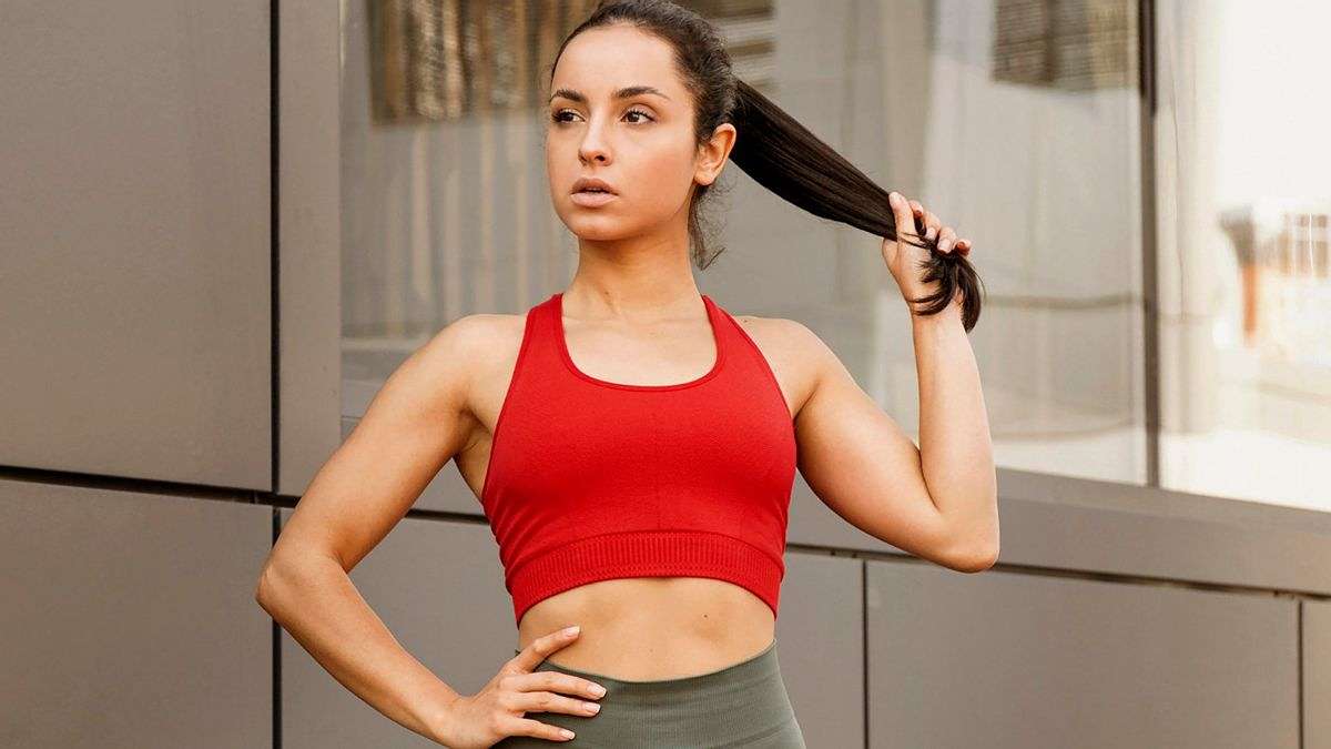 7 Rahasia Ampuh Cara Menata Rambut Agar Tidak Lepek dan Bau Setelah Olahraga 50 Seorang wanita berambut gelap memakai sport bra merah dan memegang rambutnya, berpose di luar ruangan dengan latar dinding modern