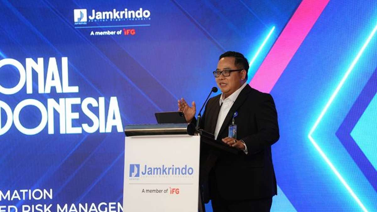 Seorang pria berkacamata berjas hitam sedang berbicara di atas podium berlogo Jamkrindo dengan latar belakang biru bernuansa perusahaan